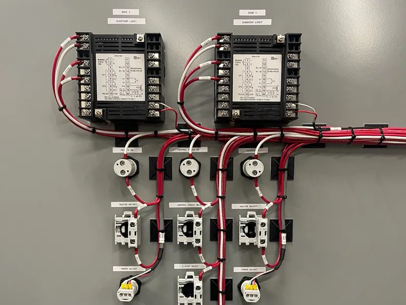The inside of a panel door: two temperature controllers labeled Zone 1 and Zone 2 overtemp limit, with pilot lights and switches below, wired in red and white with every wire numbered